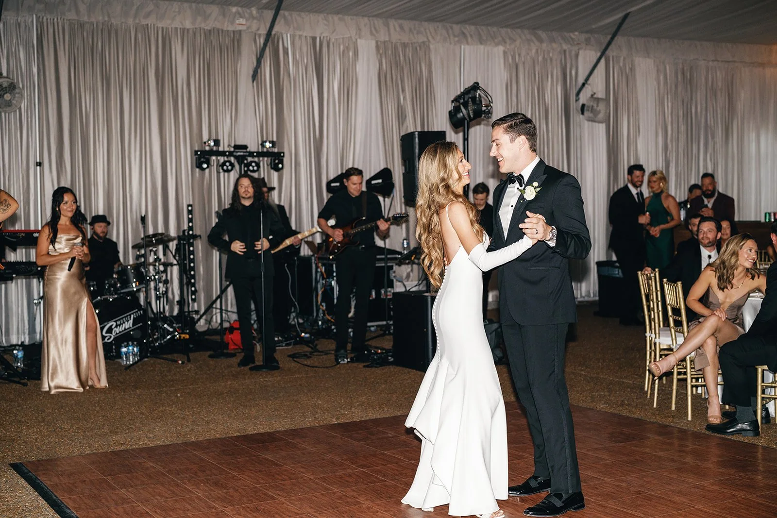 A bride and groom dancing at their wedding reception, with a live band playing in the background, surrounded by guests seated at tables.
