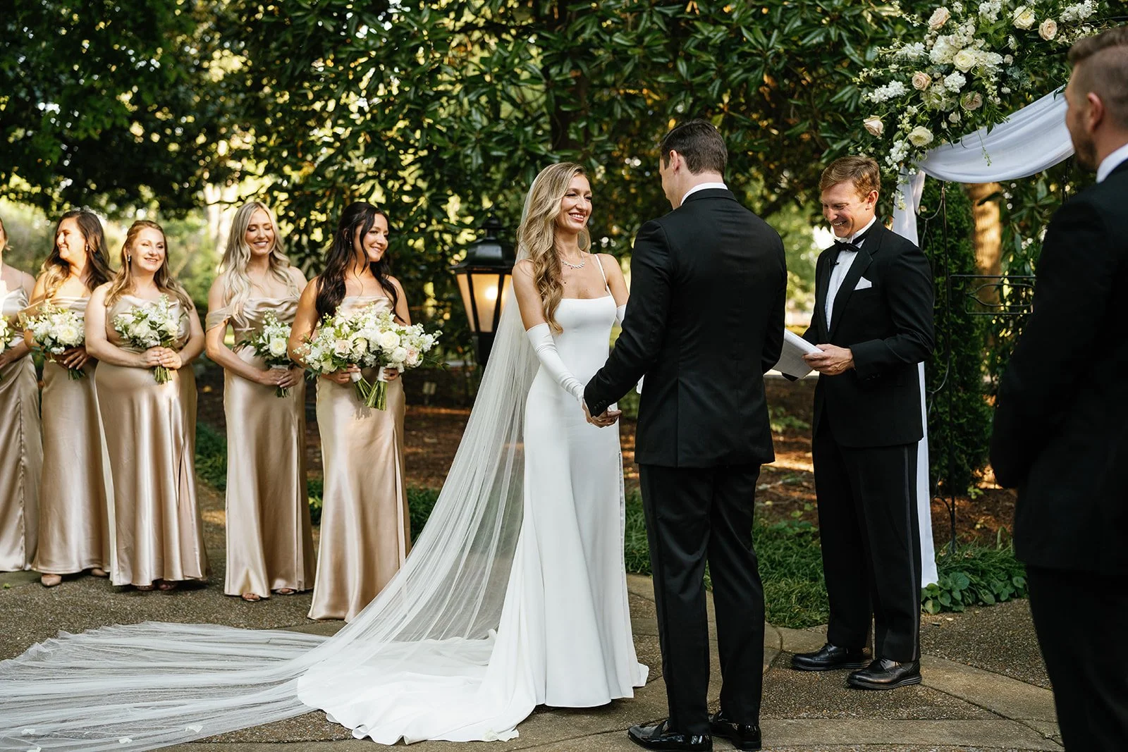 Bride and groom holding hands during outdoor wedding ceremony, with bridesmaids and groomsmen standing nearby, surrounded by greenery and floral decorations.