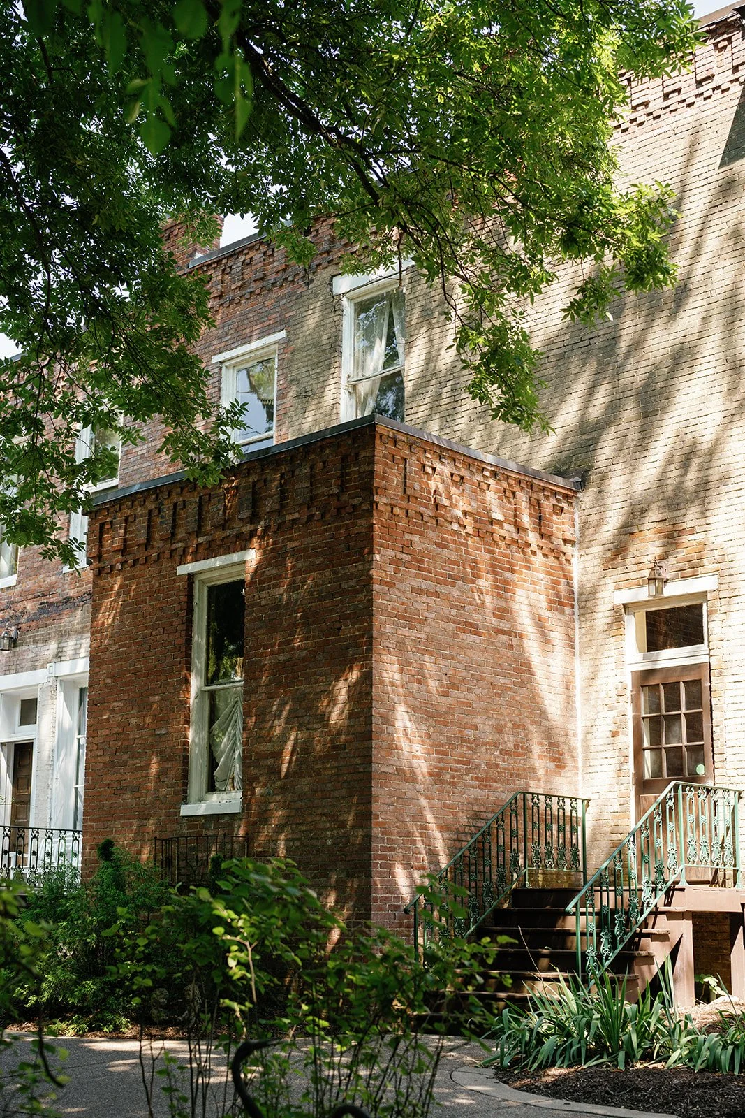 A brick townhouse with stairs leading up to a door, surrounded by green trees and plants, with sunlight casting shadows on the facade.