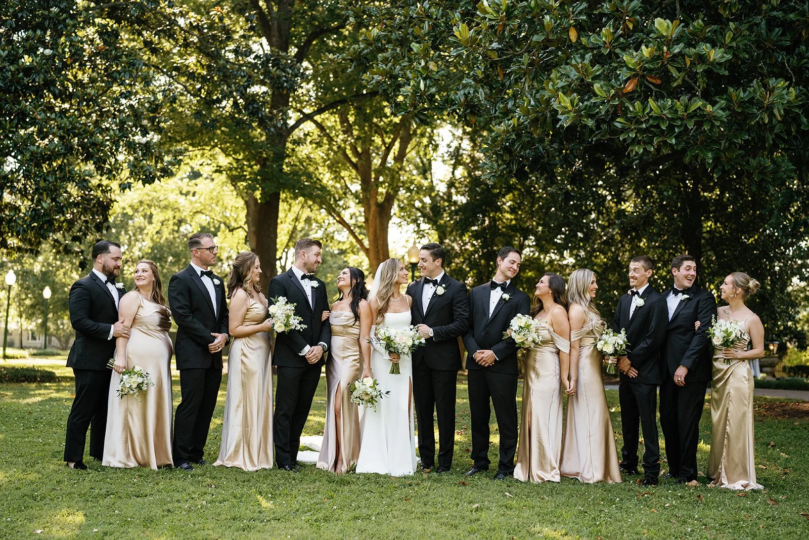 A wedding party standing outdoors on a grassy area with large trees and sunlight filtering through the leaves. The bride and groom are in the center, surrounded by bridesmaids and groomsmen, all dressed in formal attire and smiling at each other.