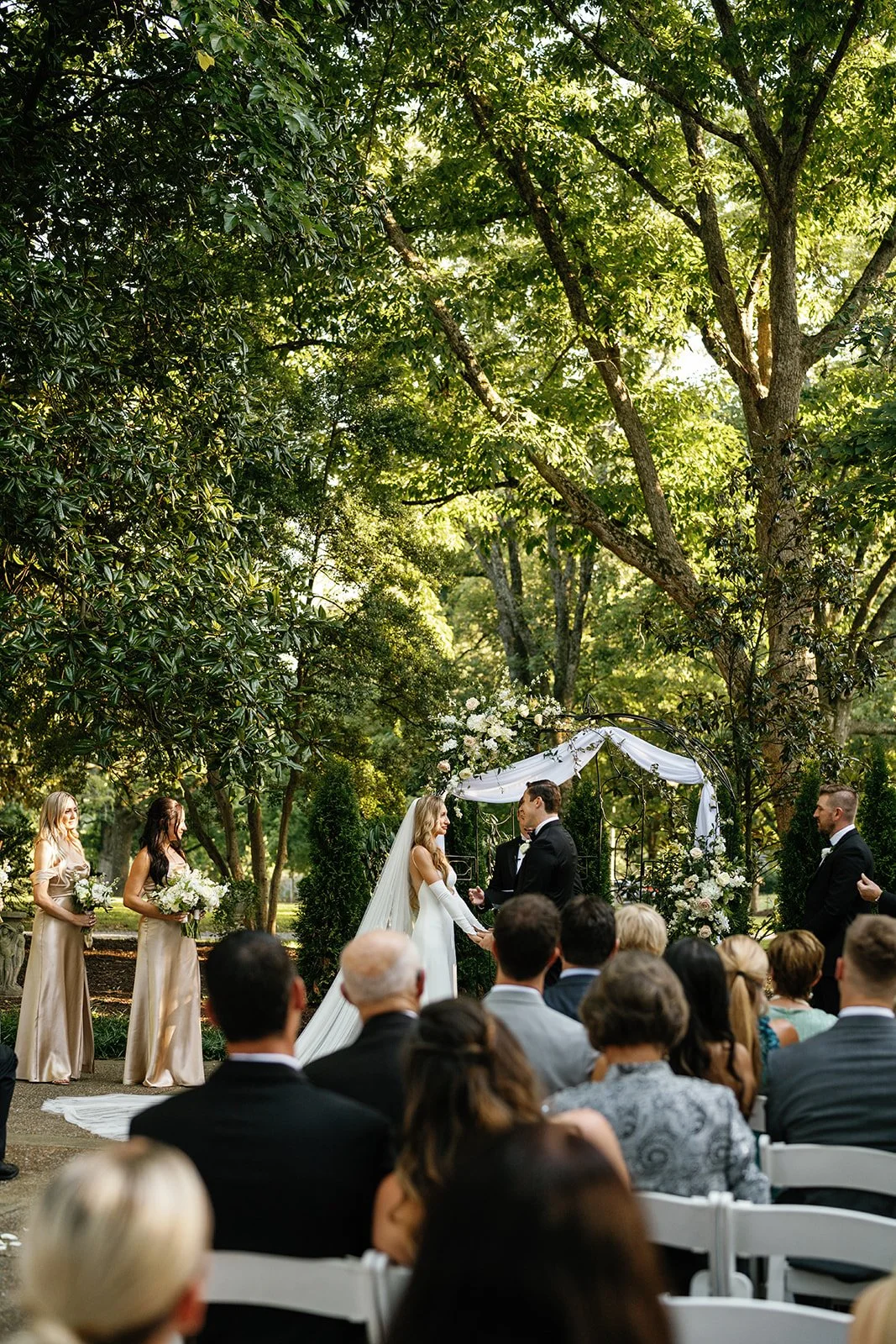 A wedding ceremony taking place outdoors in a lush green park with trees overhead. The bride and groom are holding hands under a floral archway, with bridesmaids on one side and a groomsman on the other. Guests are seated in white chairs watching the