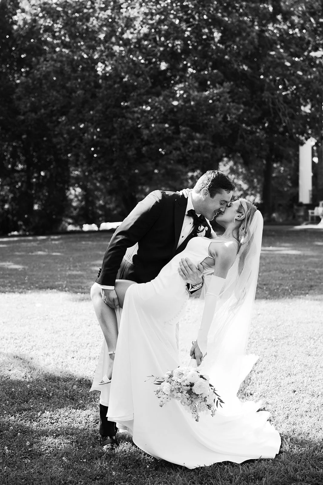 A black and white photo of a bride and groom. The groom is dipping the bride while they kiss. The bride is holding a bouquet of flowers, and they are outdoors with trees in the background.