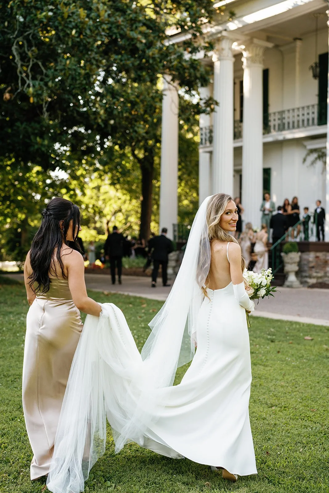A bride in a white gown holding a bouquet smiles as she walks on a lawn, assisted by a bridesmaid in a beige dress, near a white mansion with tall columns and a terrace, with guests gathered in the background.