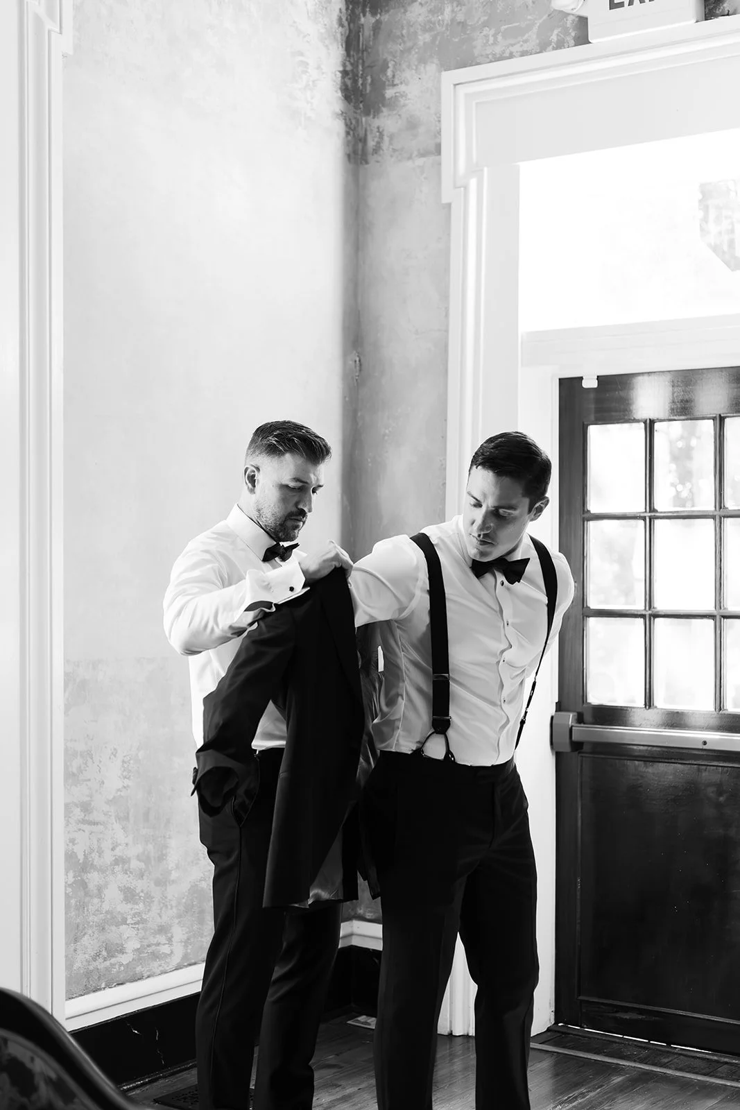 Two men in tuxedos grooming each other's suits, near a window and a door, in black and white.