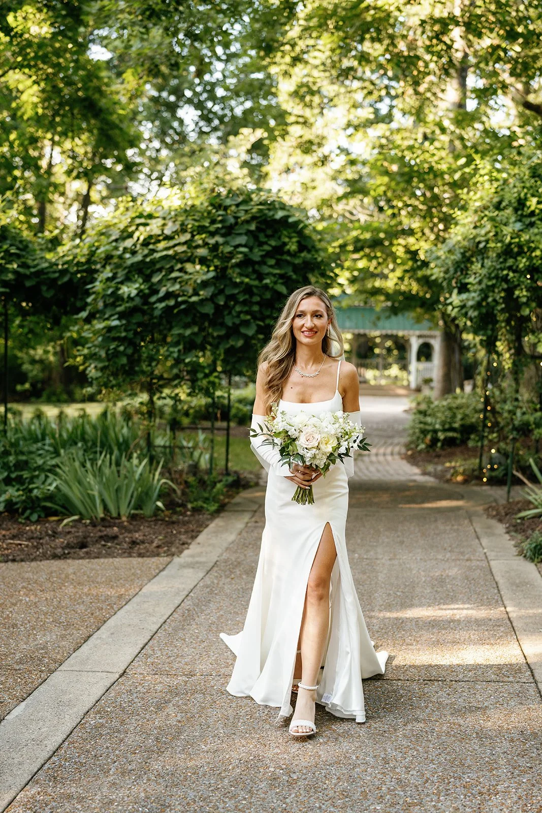 A smiling bride in a white wedding dress with a high slit, holding a bouquet of white flowers, walking on a garden pathway surrounded by lush green trees and plants.