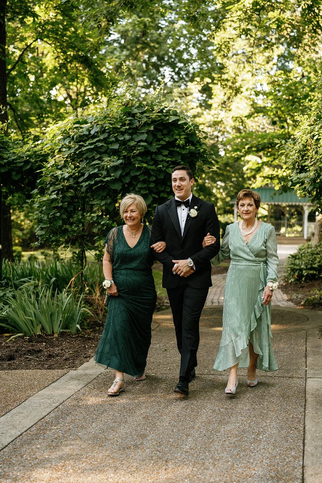 A young man in a black tuxedo walking arm-in-arm with two women in formal dresses outdoors on a garden pathway surrounded by lush green foliage.
