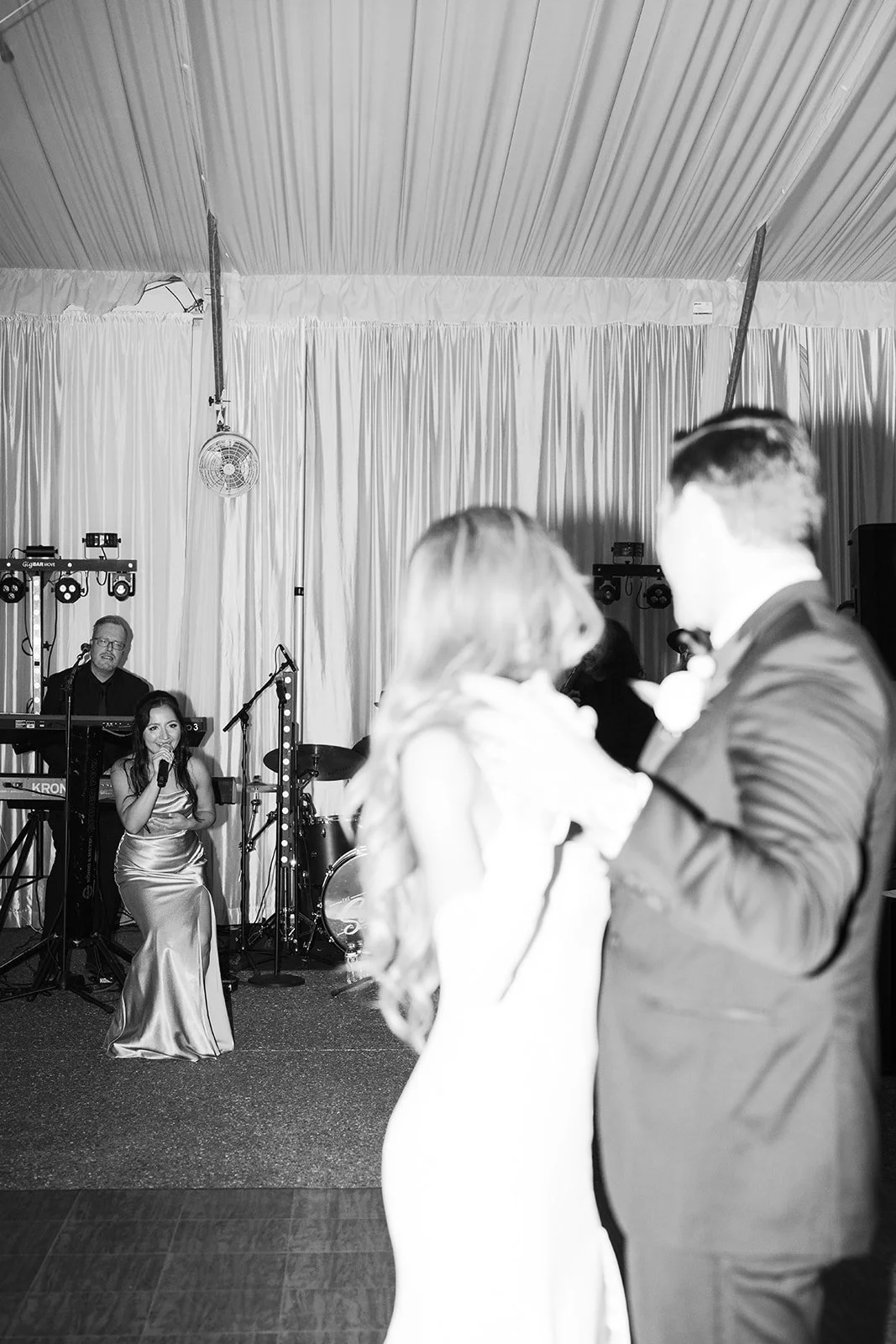 Black and white photo of a wedding reception with a bride and groom dancing, a singer sitting on the floor singing into a microphone, and a keyboardist behind them, all inside a tent with curtains and a ceiling fan.