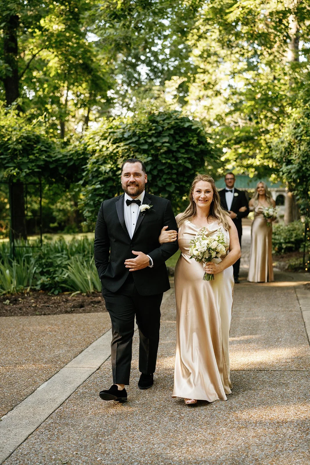 A wedding procession with a man and a woman walking arm-in-arm outdoors on a sunny day surrounded by green trees; the man is in a tuxedo with a bow tie and the woman is in a champagne-colored satin dress holding a bouquet of white and cream flowers.