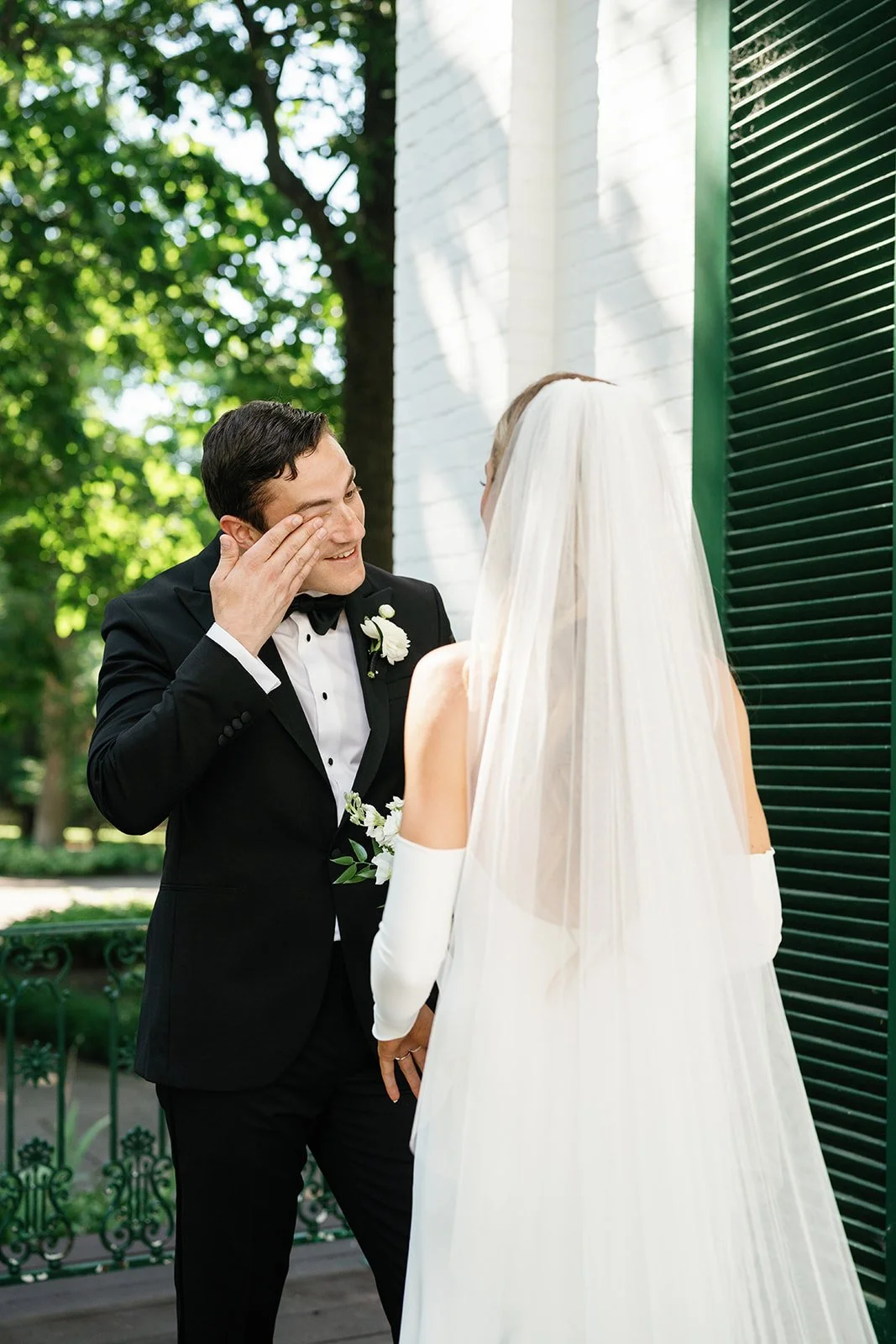 A groom in a black tuxedo with a white boutonniere wiping tears of joy as a bride in a white wedding dress and veil looks at him during their outdoor wedding ceremony.