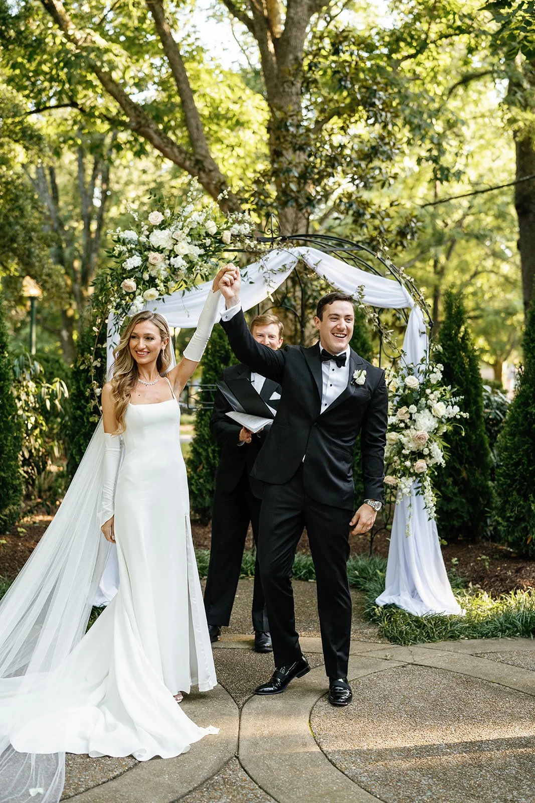 A newly married couple holding hands and smiling at their wedding ceremony outdoors, under a floral arch with greenery and white flowers.