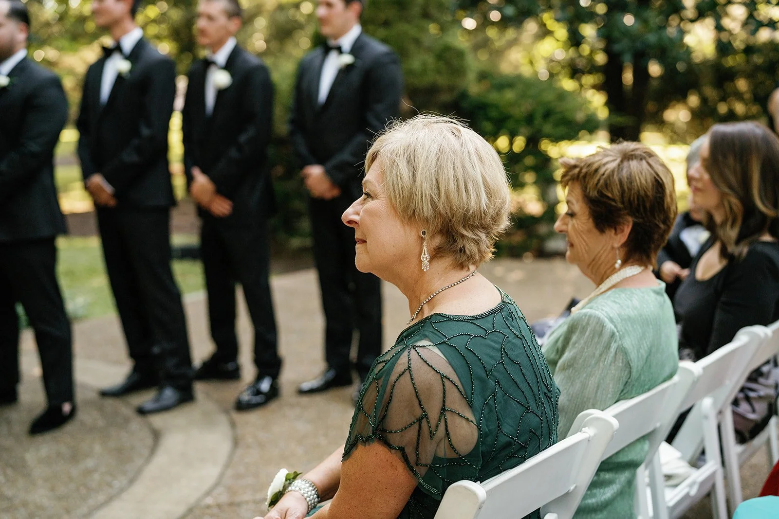 Women seated outdoors attending a formal event, with men in tuxedos standing in the background.