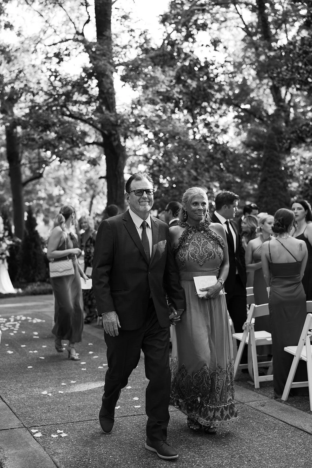 Black and white photo of a man and woman walking hand in hand at an outdoor event, with other people socializing in the background under trees.