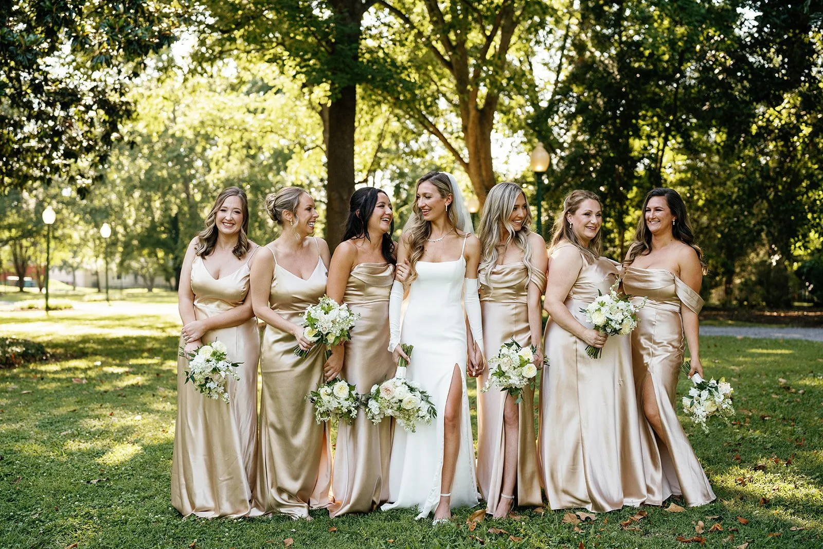 A bride in a white wedding dress with a thigh-high slit and six bridesmaids in matching champagne-colored satin dresses holding bouquets, standing outdoors in a park with trees and sunlight.
