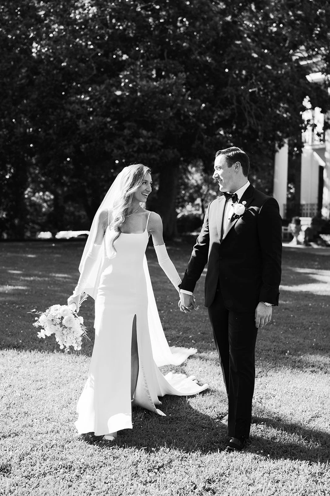 A bride and groom holding hands and smiling at each other outdoors, with trees and a building in the background, in black and white.