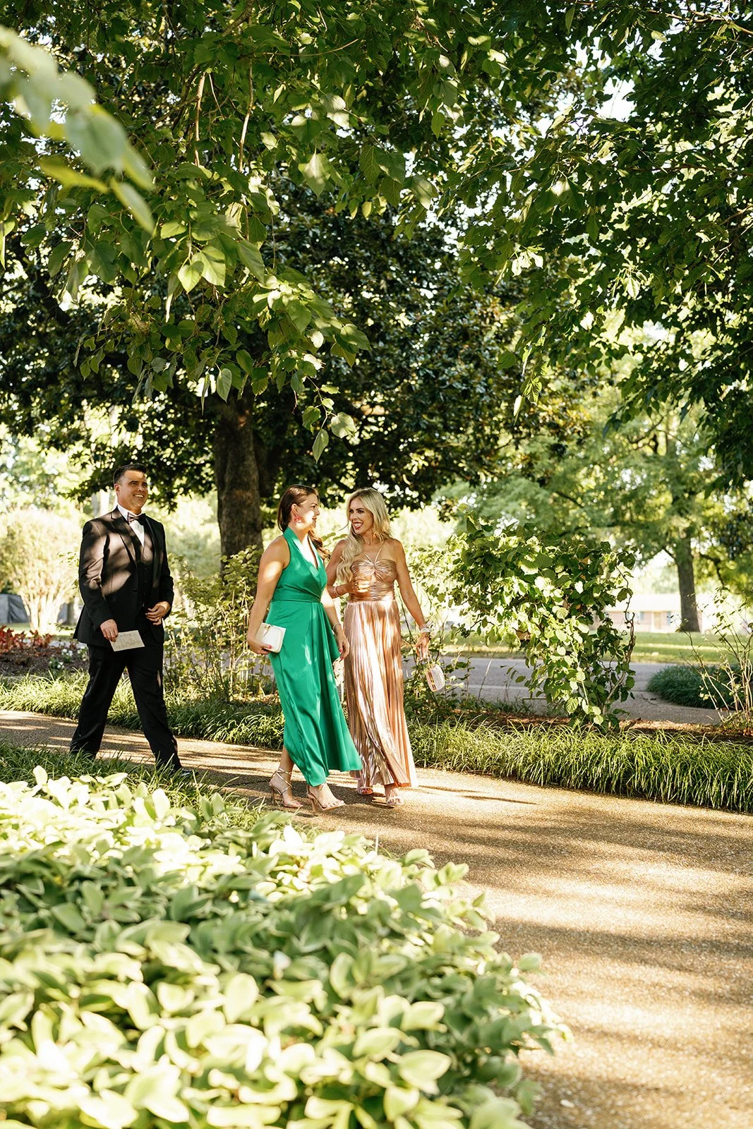Three people dressed in formal attire walking and conversing on a sunny garden pathway surrounded by lush green trees and bushes.