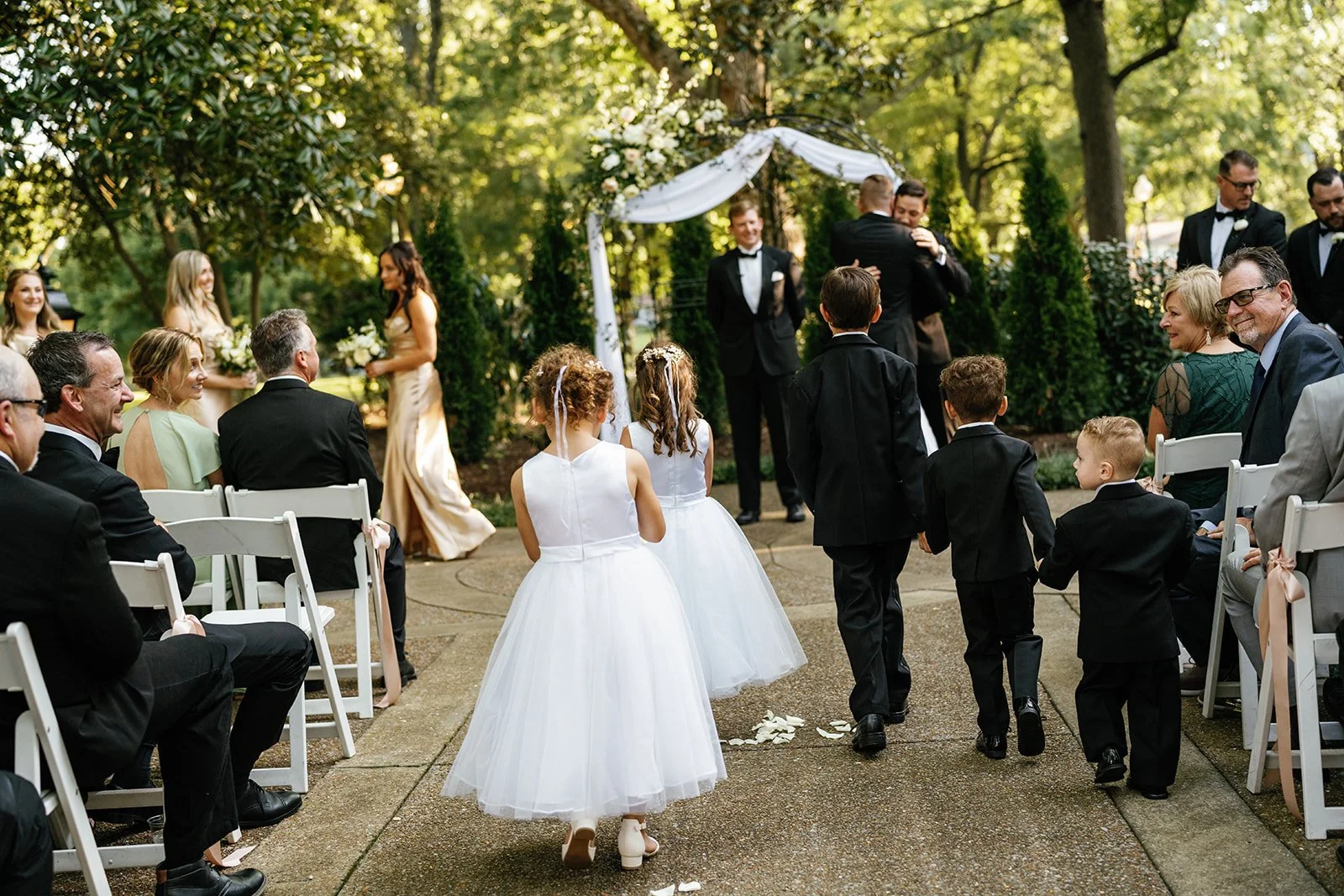 Children and adults attending an outdoor wedding ceremony, with kids walking down the aisle dressed in formal clothes, surrounded by seated guests and greenery.