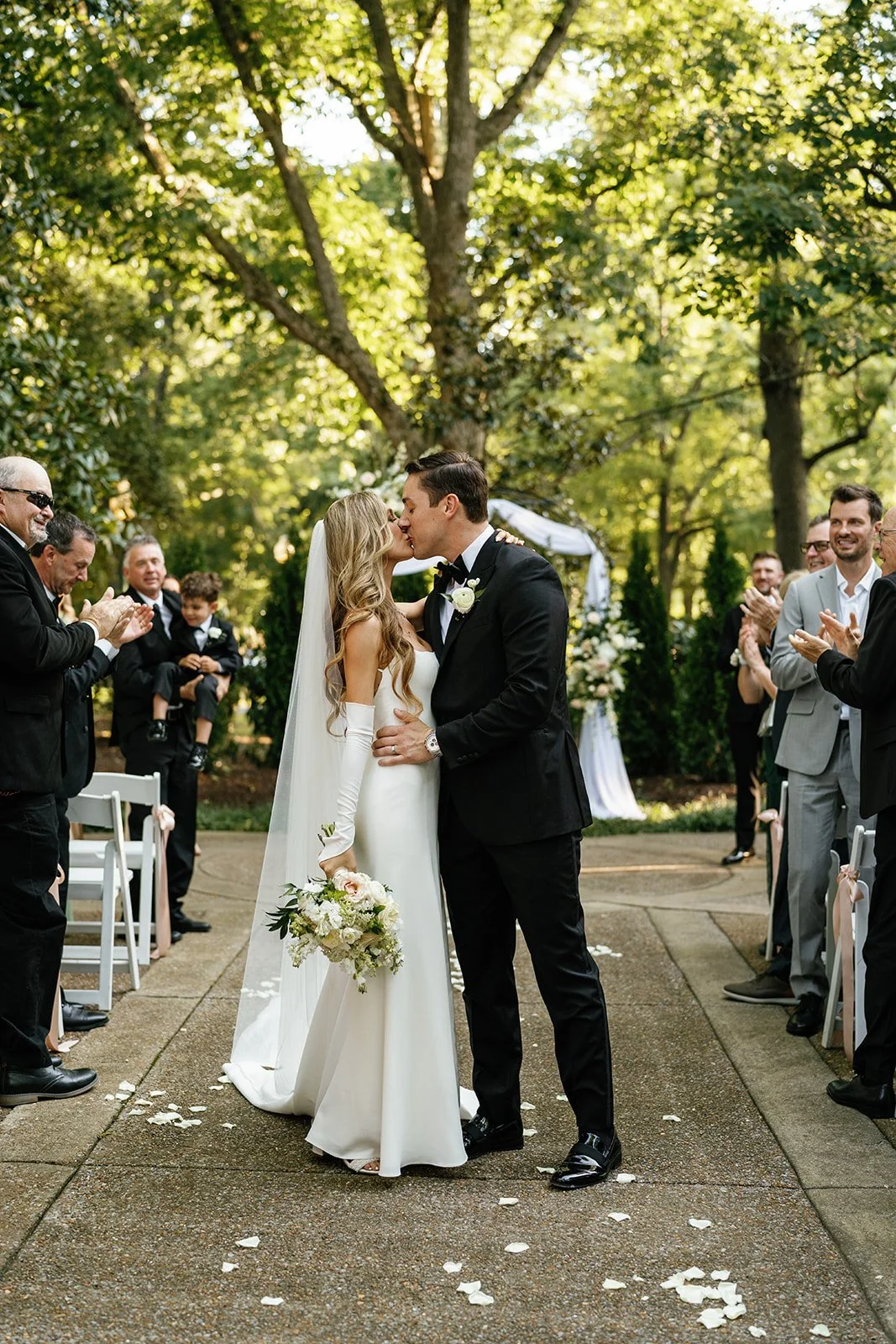 A bride and groom share a kiss under a arbor at an outdoor wedding ceremony with guests clapping and celebrating around them.