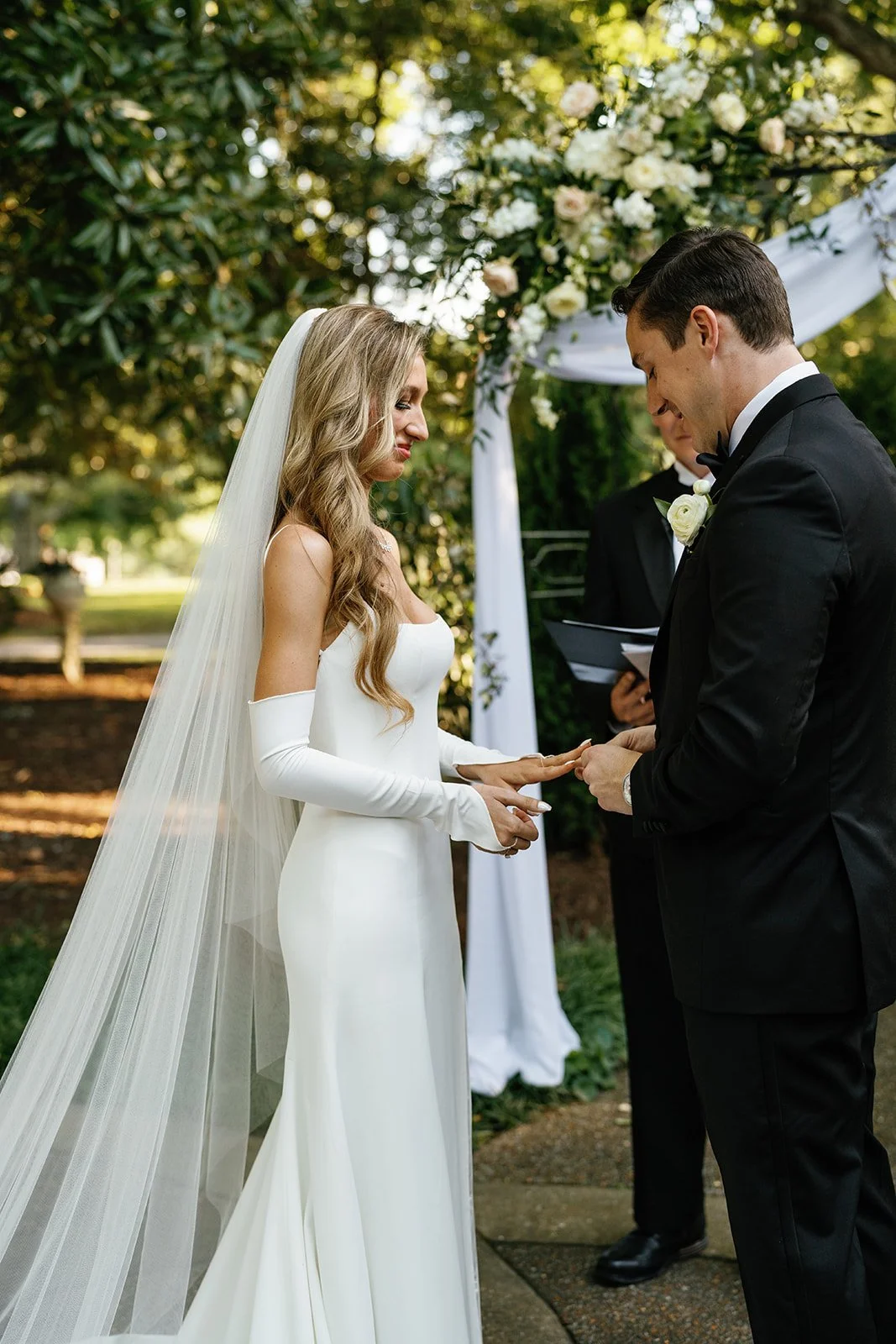 Bride and groom exchange rings during outdoor wedding ceremony under floral arch.