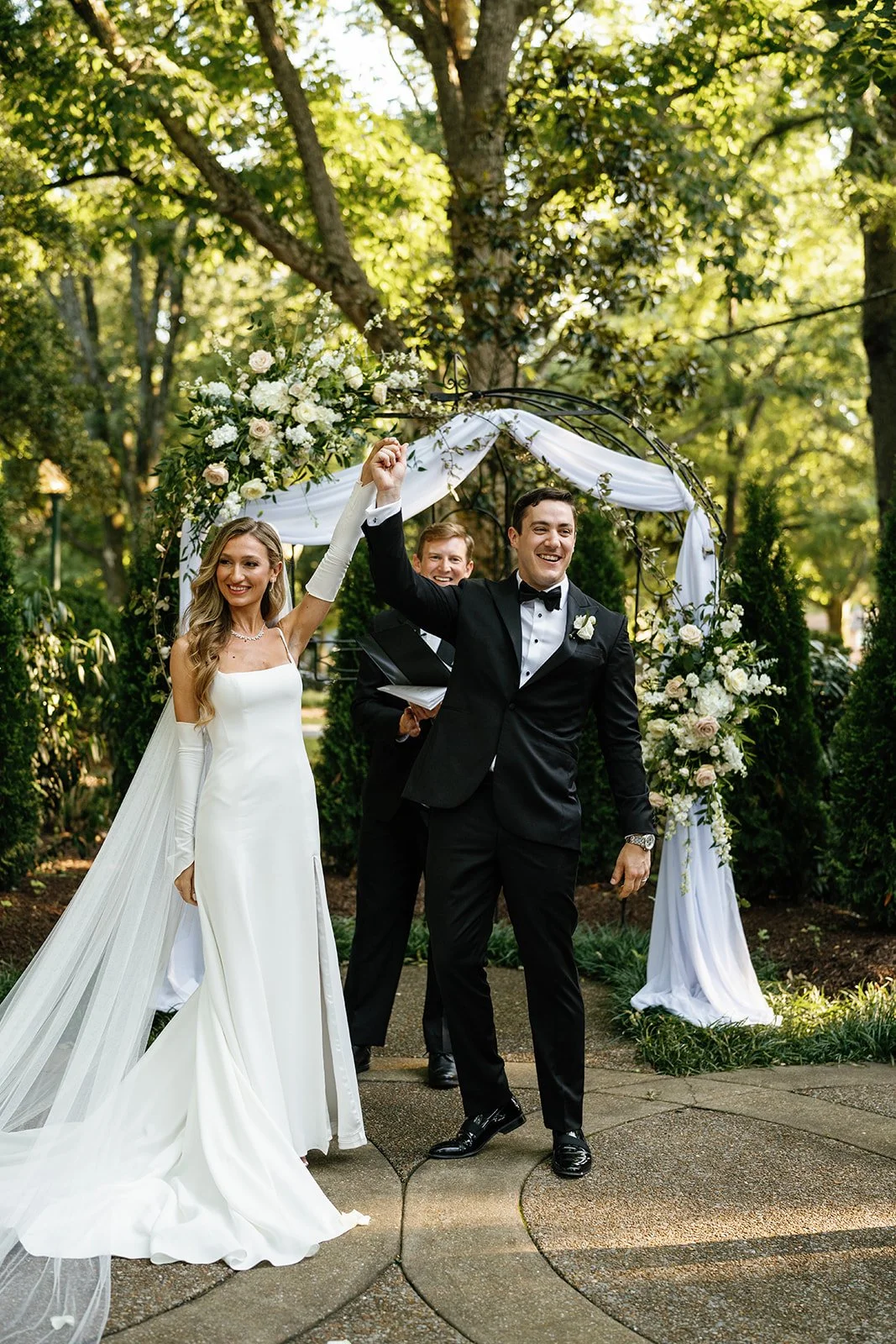 A newly married couple celebrating under a decorated wedding arch outdoors surrounded by trees. The bride is in a white wedding gown with a long train and veil, and the groom is in a black tuxedo. They are holding hands up in victory, smiling joyfull