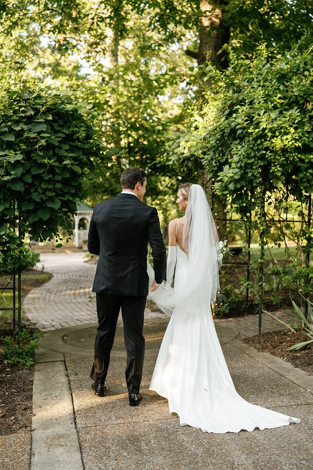 Bride and groom walking hand in hand on a garden path, surrounded by lush green trees and bushes, framed by an archway.