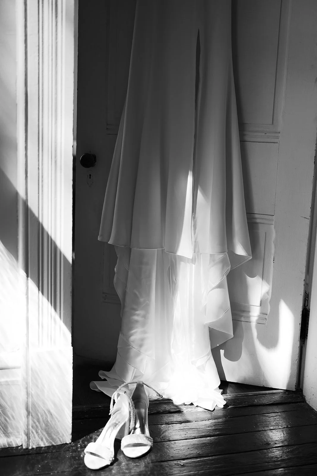 A wedding dress and high-heeled shoes hanging in a doorway, illuminated by sunlight.