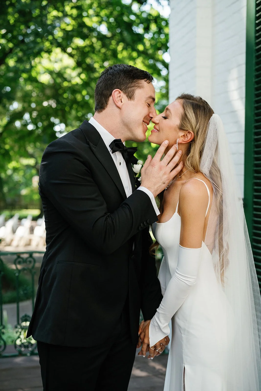 A bride and groom on their wedding day, standing outdoors, leaning in with their noses and foreheads touching, smiling with eyes closed, holding hands, with trees and a white building in the background.
