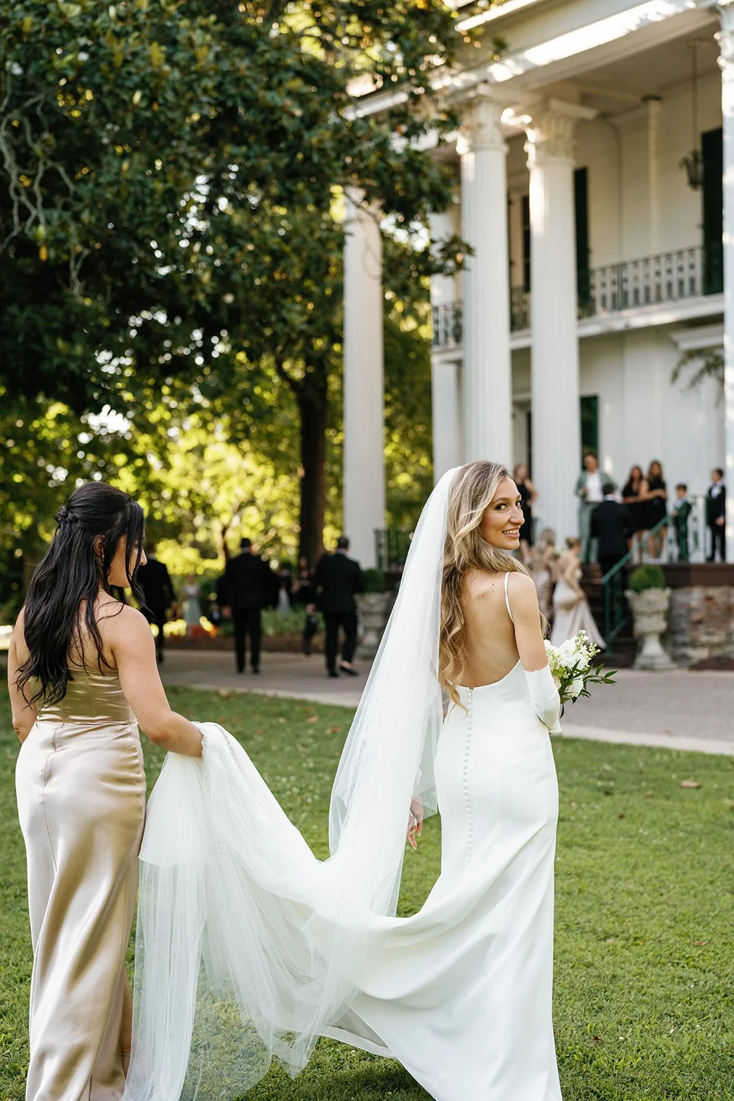 Bride in a white wedding gown and veil holding a bouquet, smiling, in a garden with a large white house and bridesmaids in the background.