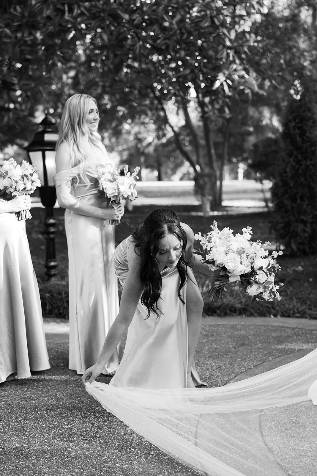A bride and bridesmaids preparing for a wedding outdoors, with one bridesmaid adjusting her gown while holding a bouquet, and other bridesmaids standing nearby holding flowers.