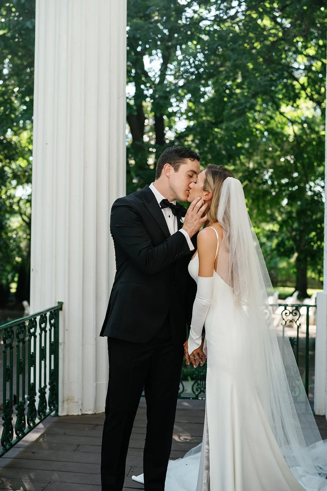 A bride and groom sharing a kiss outdoors, holding hands, with green trees in the background and a white column nearby.