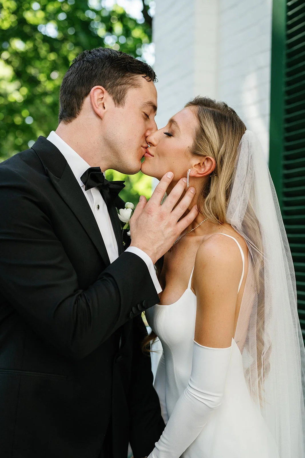 A bride and groom kissing outdoors, the groom in a black tuxedo and the bride in a white wedding dress with a veil.