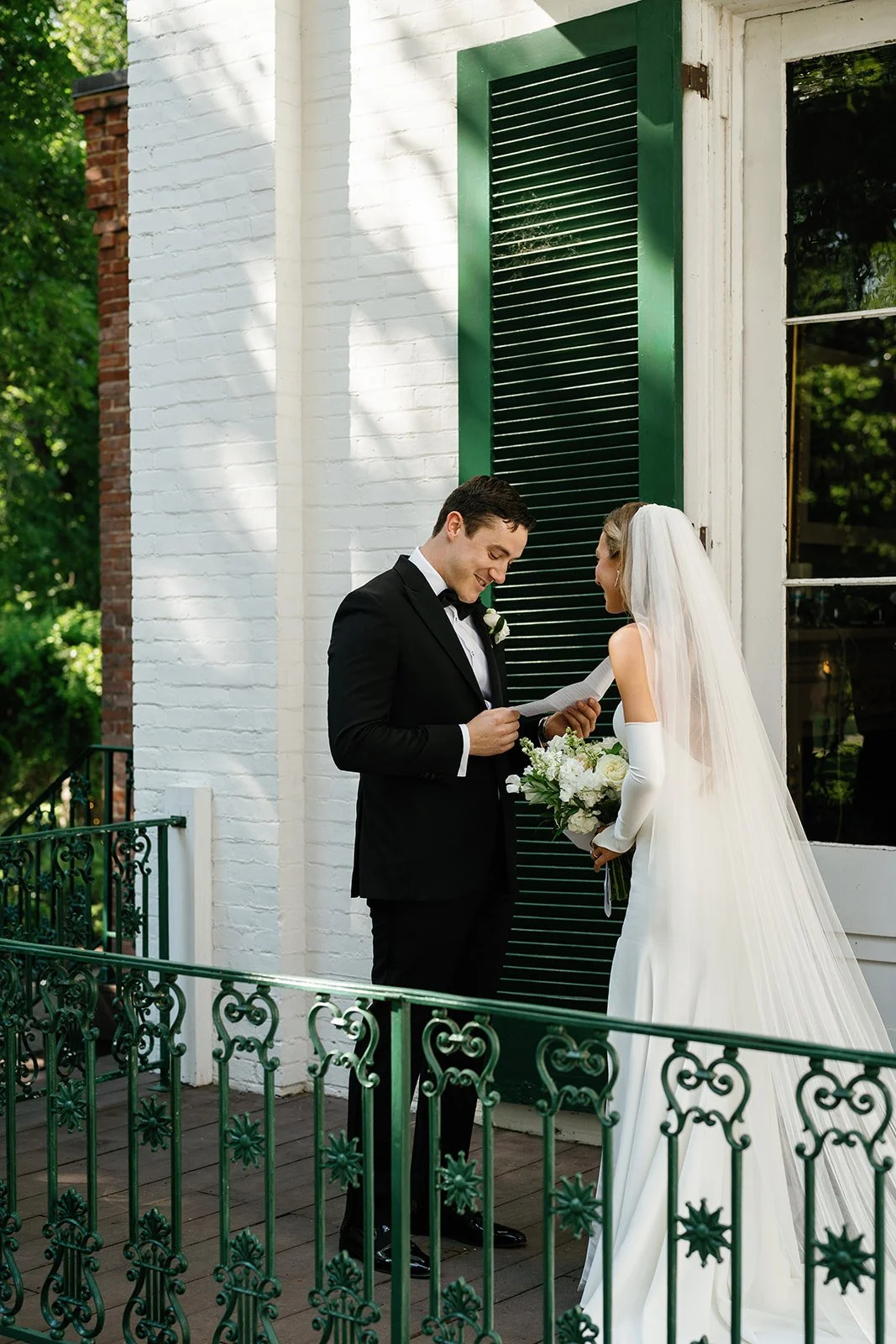 A bride and groom stand outside a white brick building with green shutters, sharing a moment during their wedding ceremony. The groom is in a black tuxedo, and the bride in a long white dress with a veil, holding a bouquet of white flowers.