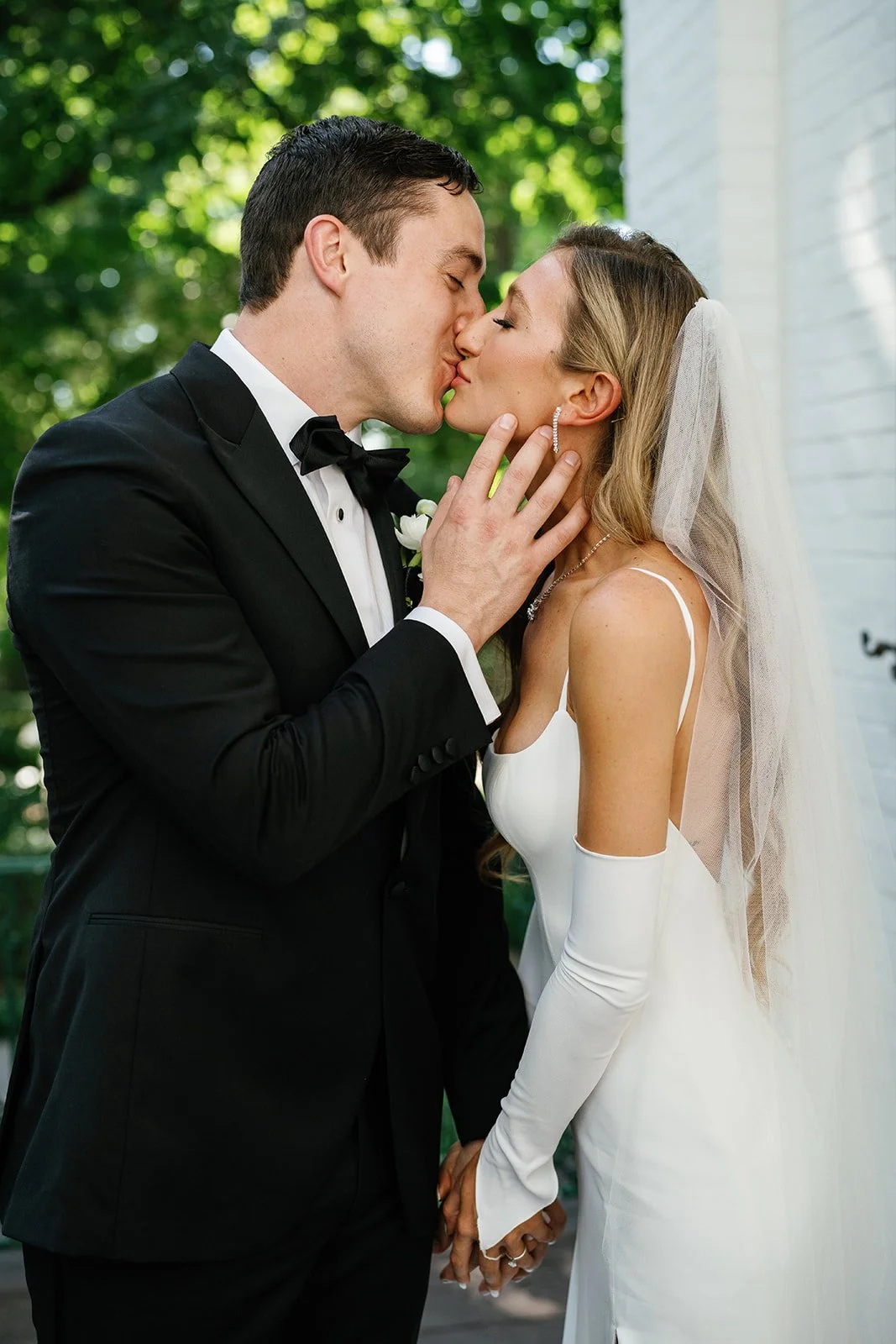 A bride and groom sharing a kiss outdoors, holding hands, with lush green trees in the background. The groom is dressed in a black tuxedo with a bow tie, and the bride is wearing a white wedding dress with long gloves and a veil.