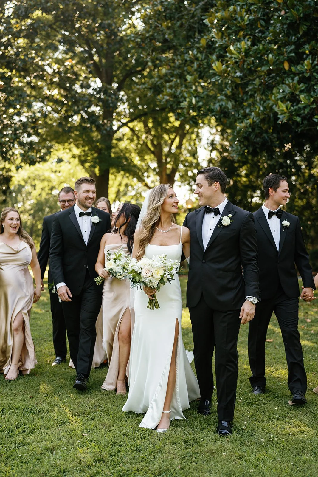 A group of wedding guests, including a bride and groom, walking outdoors on a sunny day with green trees in the background.