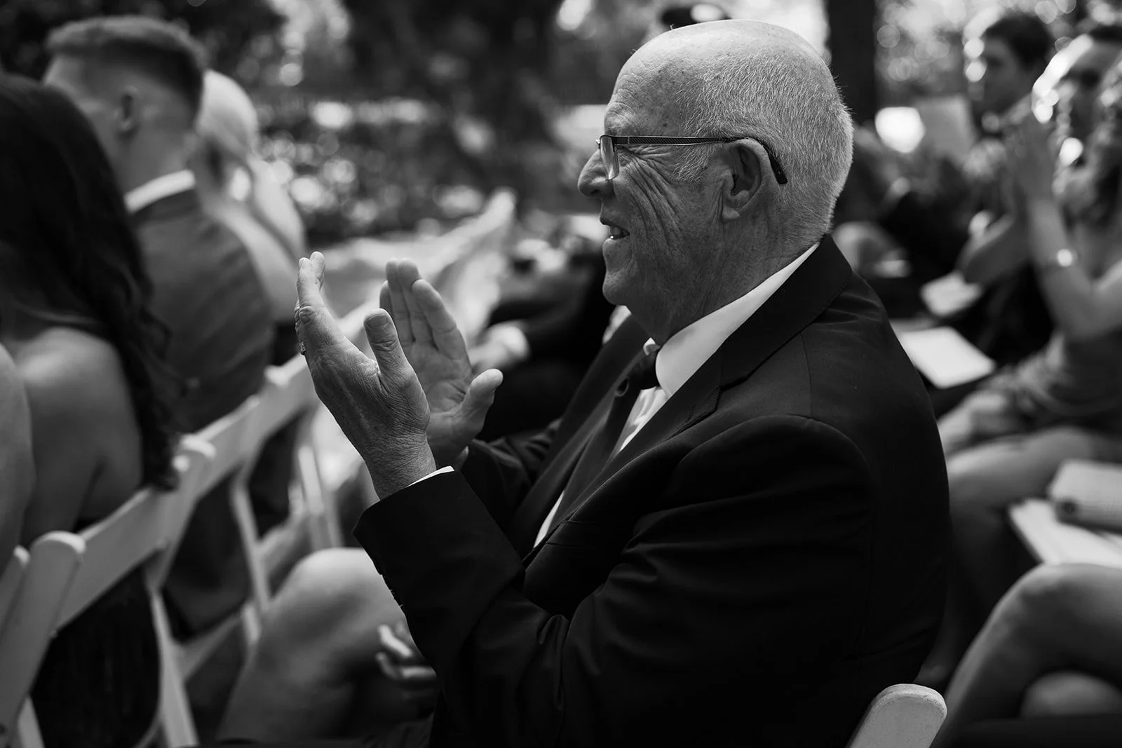 A smiling elderly man in a black suit and glasses engaging in conversation at an outdoor event.
