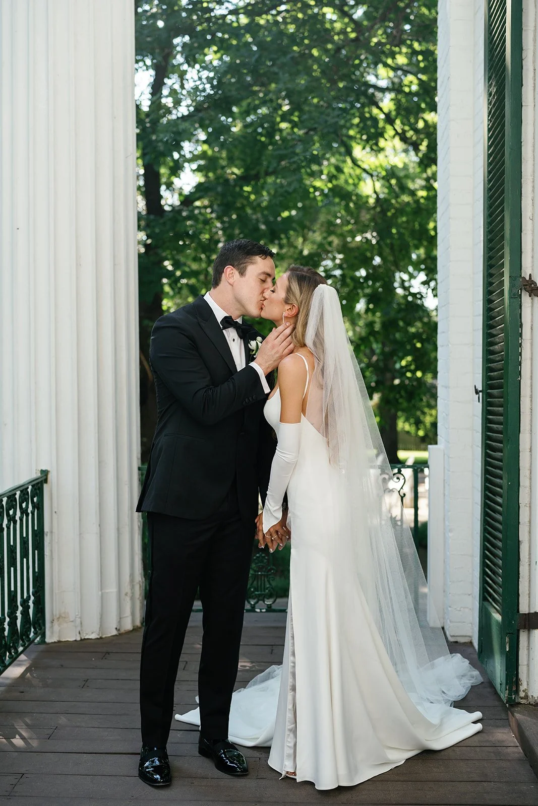 A newlywed couple sharing a kiss outdoors on a porch, with the groom in a black tuxedo and the bride in a white wedding gown and veil, surrounded by greenery.