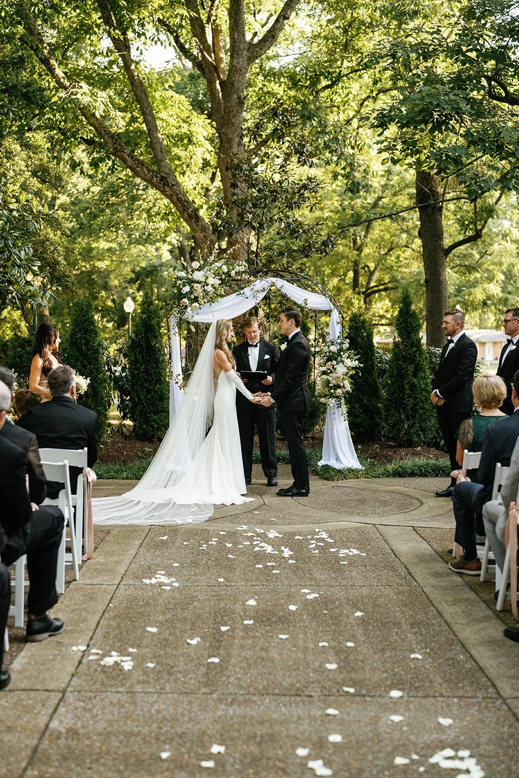 A wedding ceremony taking place outdoors in a wooded area, with the bride and groom exchanging vows under a decorative arch adorned with white flowers and drapery. Guests are seated on either side, and the ground is sprinkled with white flower petals