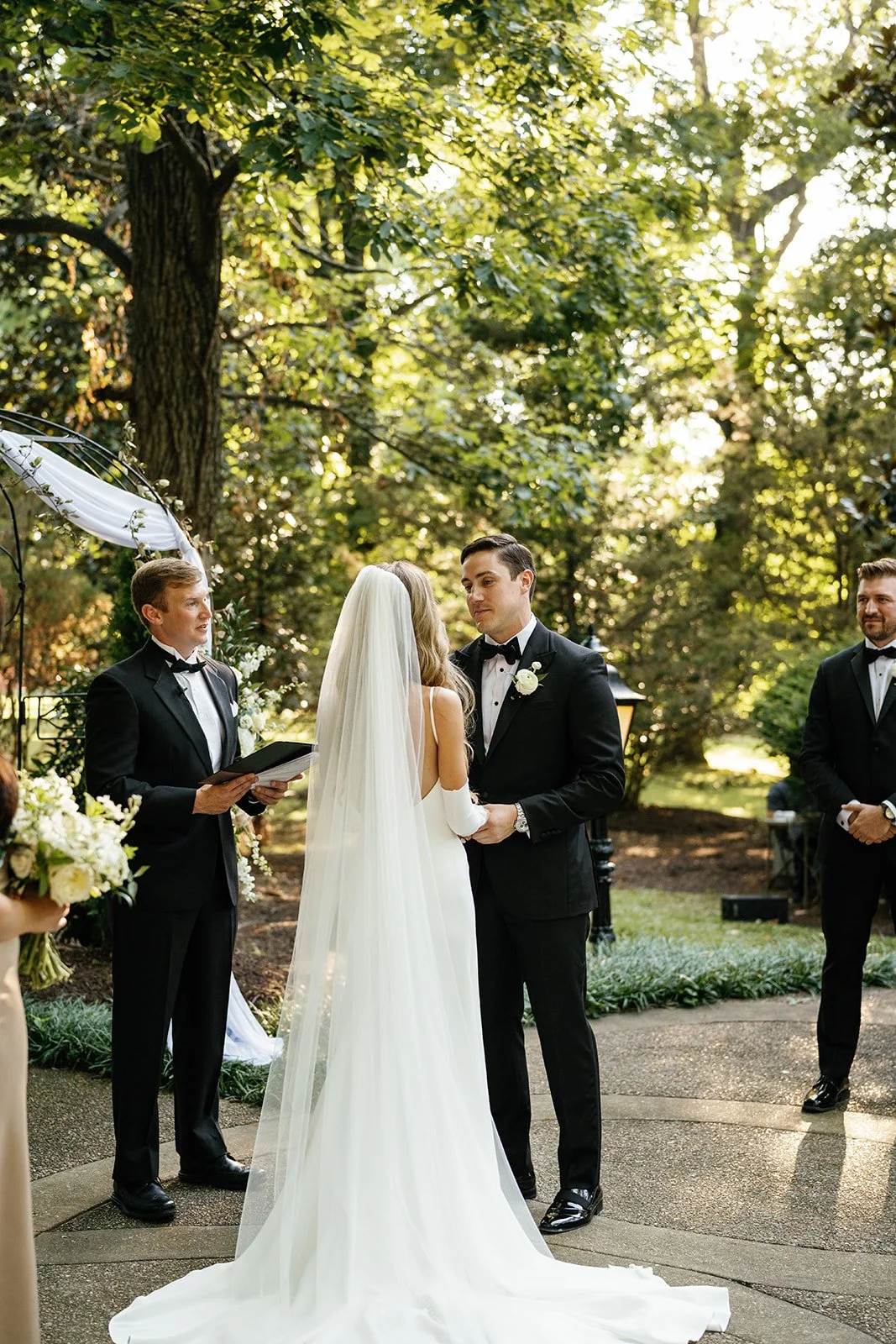 A bride and groom exchanging vows at an outdoor wedding ceremony surrounded by greenery, with an officiant and groomsman present.