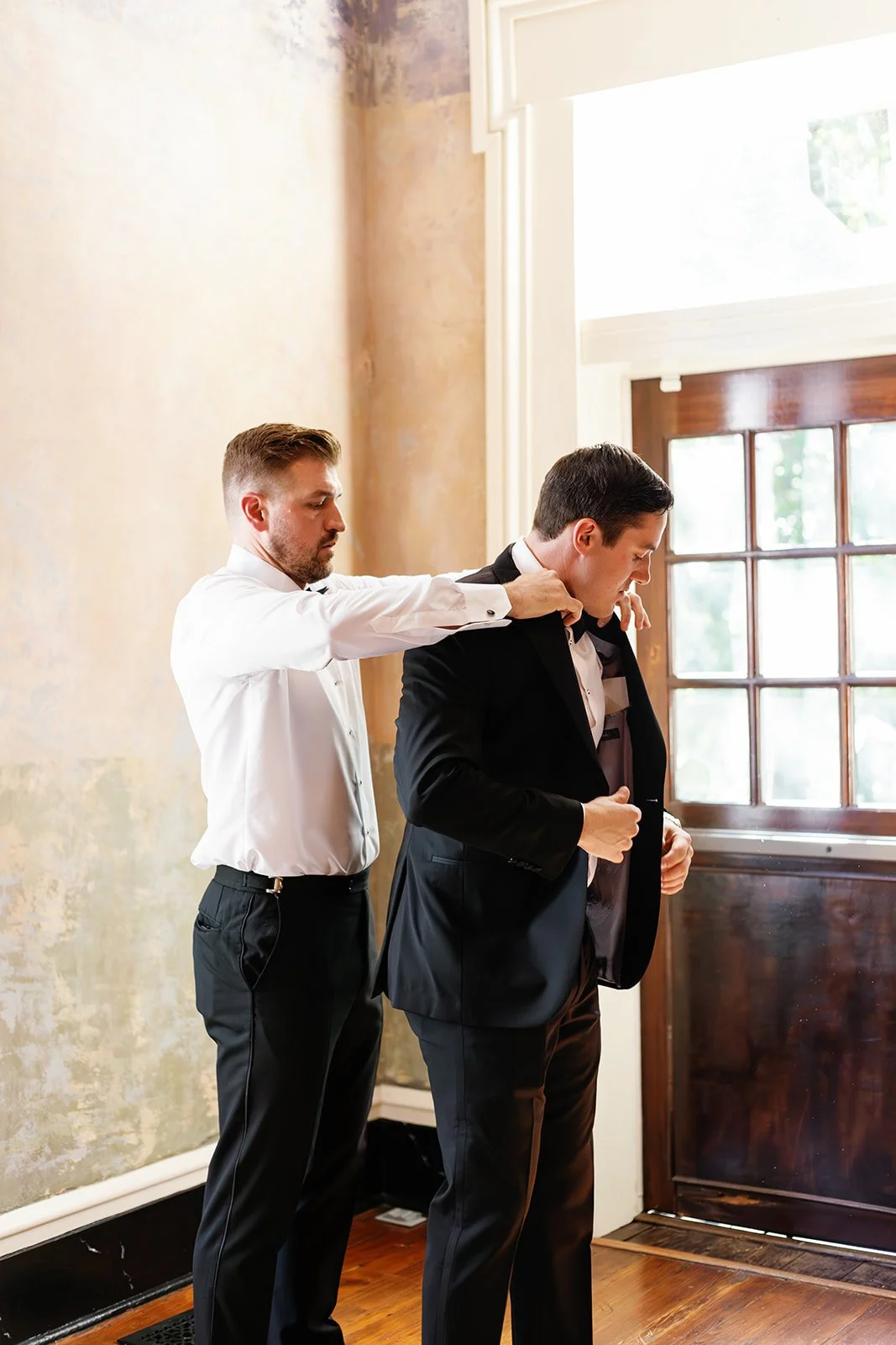 Two men in tuxedos, one helping the other adjust his jacket near a window in a warmly lit room.