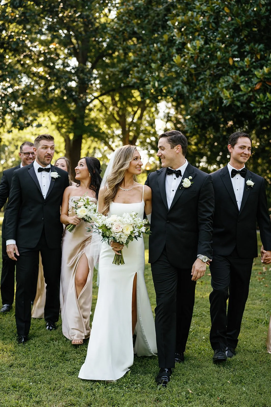 Bride and groom walking outdoors with wedding party, surrounded by trees, smiling at each other, woman holding a bouquet, women with dresses and men in tuxedos.