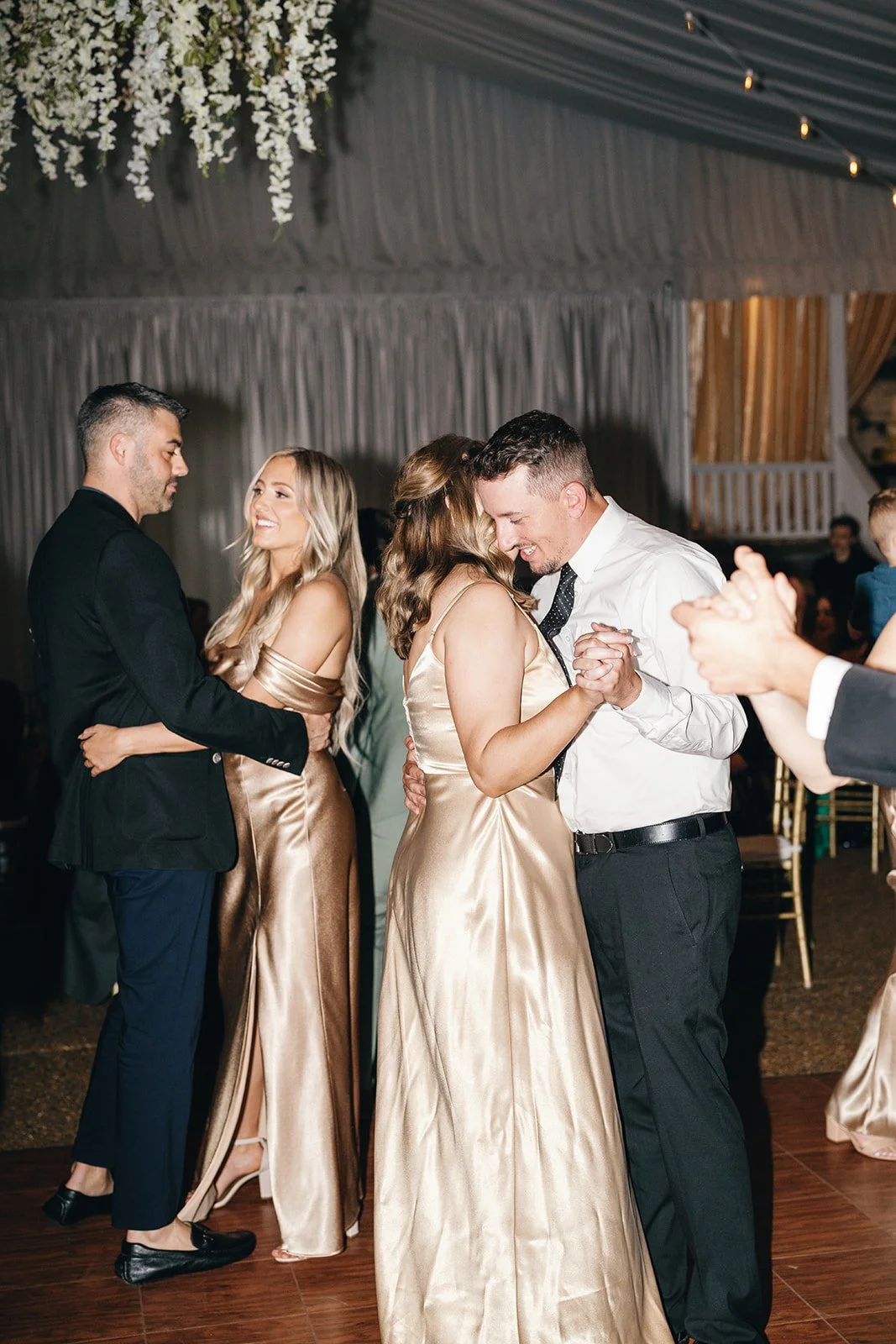 Couples dancing at a wedding reception, with two couples in focus, the background decorated with draped fabric and hanging flowers.
