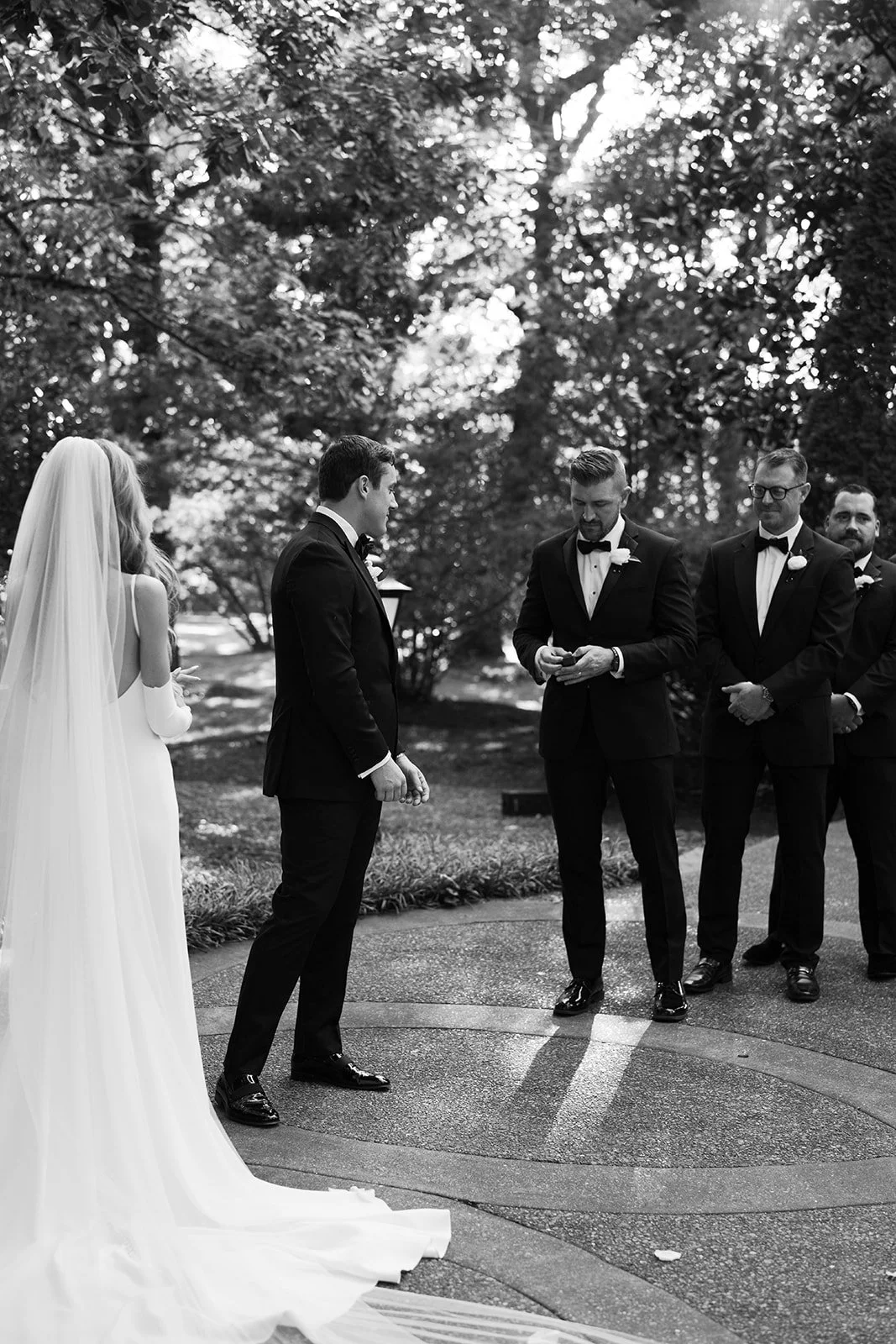 A black and white photo of a wedding ceremony outdoors with five people, including a bride in a long white gown with a veil, and four men in tuxedos, standing on a paved path with trees in the background.