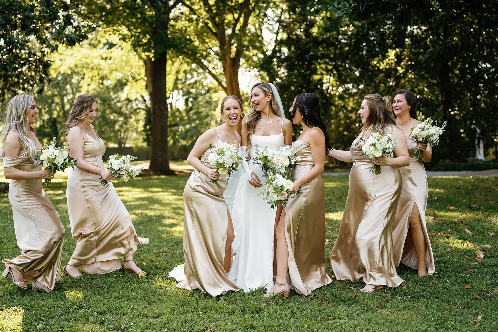 Group of women in a park, including a bride in a white wedding dress with a veil, surrounded by bridesmaids in matching gold satin dresses. They are all holding bouquets of white flowers and appear to be celebrating joyfully.