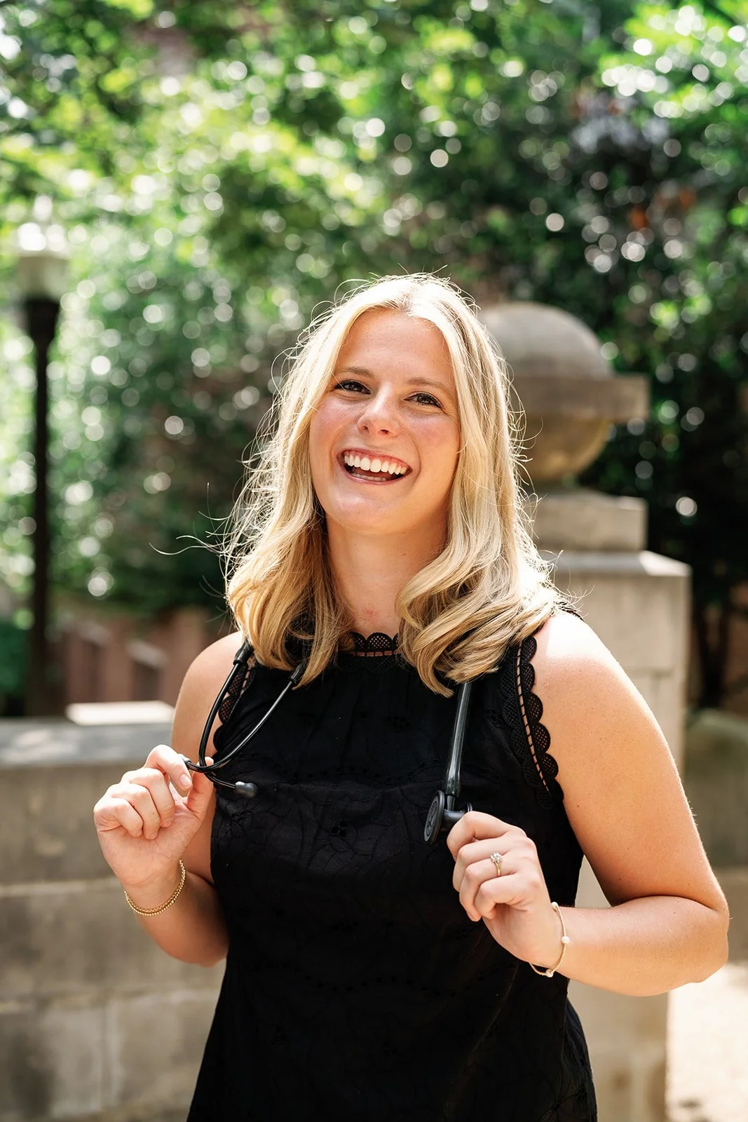 A young woman with blonde hair smiling outdoors, holding a stethoscope, outside of Vanderbilt Campus.