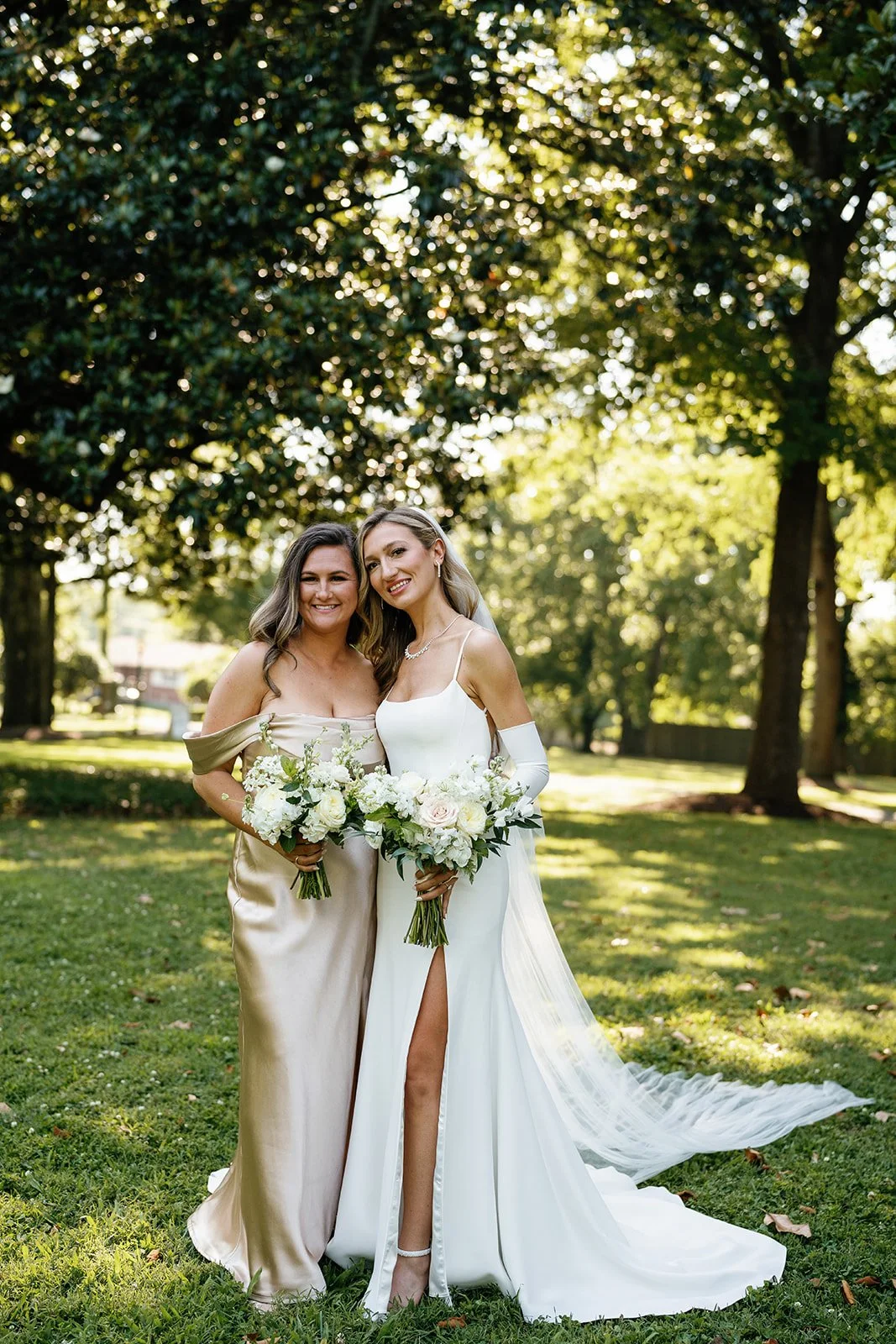 Two women in wedding attire standing outdoors at a park, smiling and holding bouquets of white flowers, with trees and sunlight in the background.