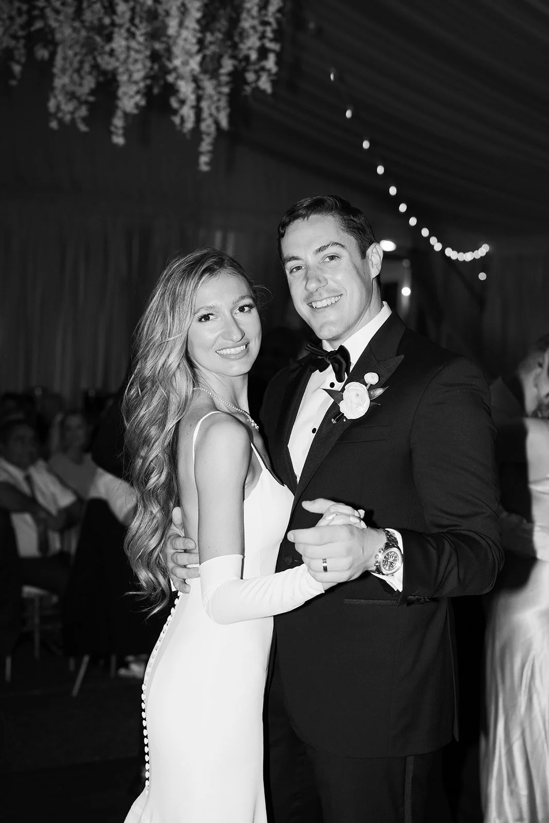 A black and white photo of a newlywed couple dancing at their wedding reception, with the bride in a white dress and long gloves, and the groom in a tuxedo with a bow tie and boutonniere, surrounded by guests.