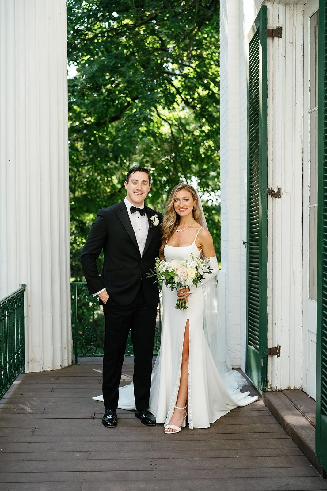A newlywed couple standing on a wooden balcony with white walls and green shutters, surrounded by lush green trees. The groom is dressed in a black tuxedo with a bow tie, and the bride is wearing a white wedding gown with a thigh-high slit, holding a