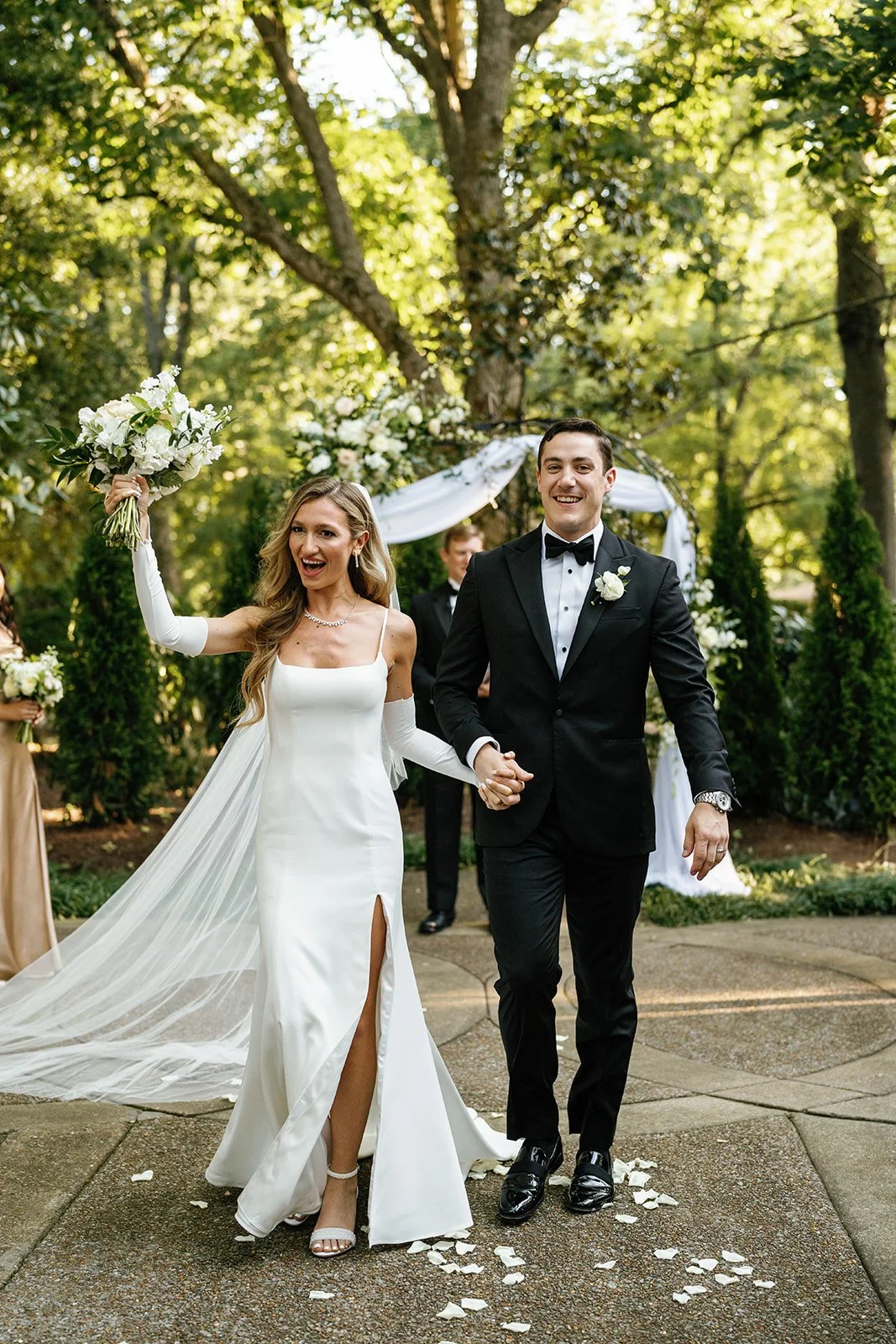 A bride and groom walking hand in hand outdoors after their wedding ceremony, with the bride holding a bouquet and the groom smiling in a black suit with a bow tie, surrounded by greenery.