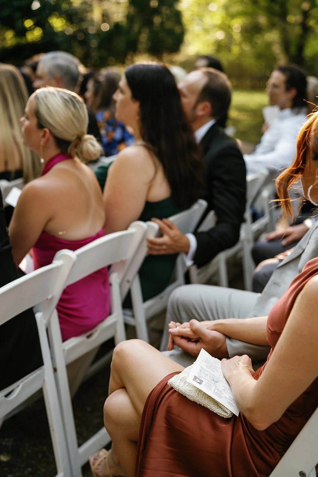 Guests seated outdoors at an event, some holding programs, with trees and sunlight in the background.