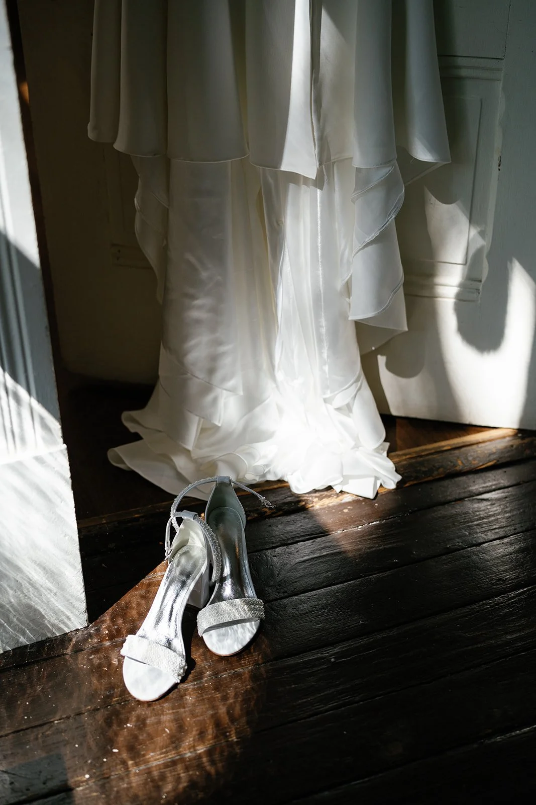 A white wedding dress hanging with a pair of silver and white high-heeled shoes on a wooden floor.