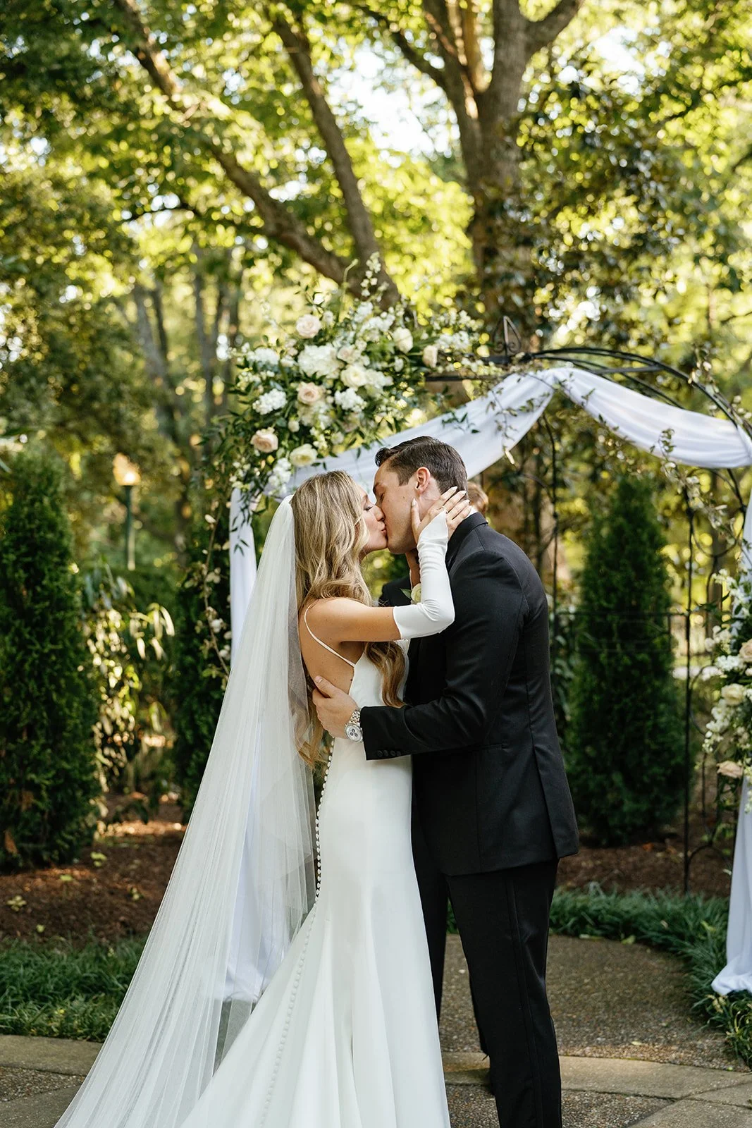 A bride and groom share a kiss during their outdoor wedding ceremony under a decorated archway with flowers and white fabric, surrounded by lush greenery and tall trees.